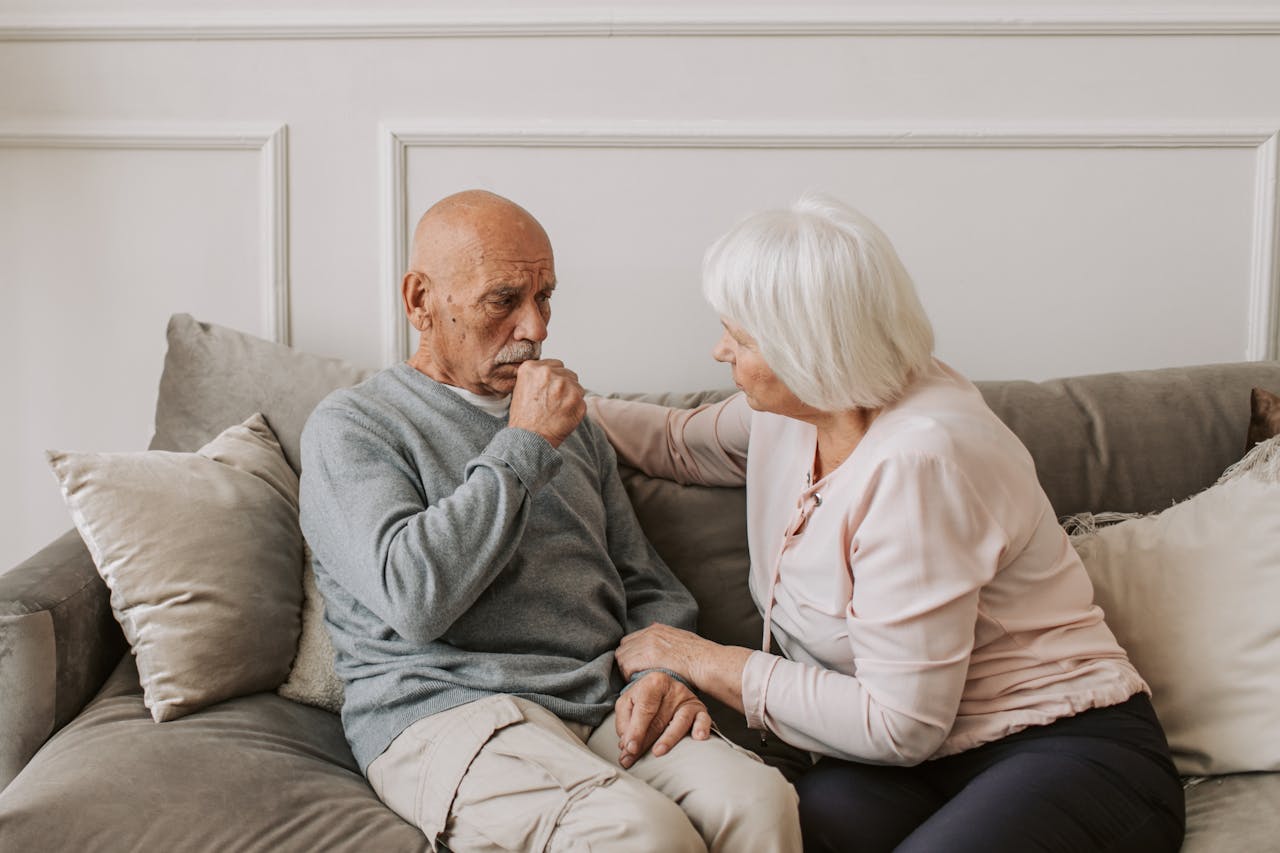 unsere Zusammenarbeit mit Polen An elderly woman comforts a man coughing on a couch, showcasing care and affection.