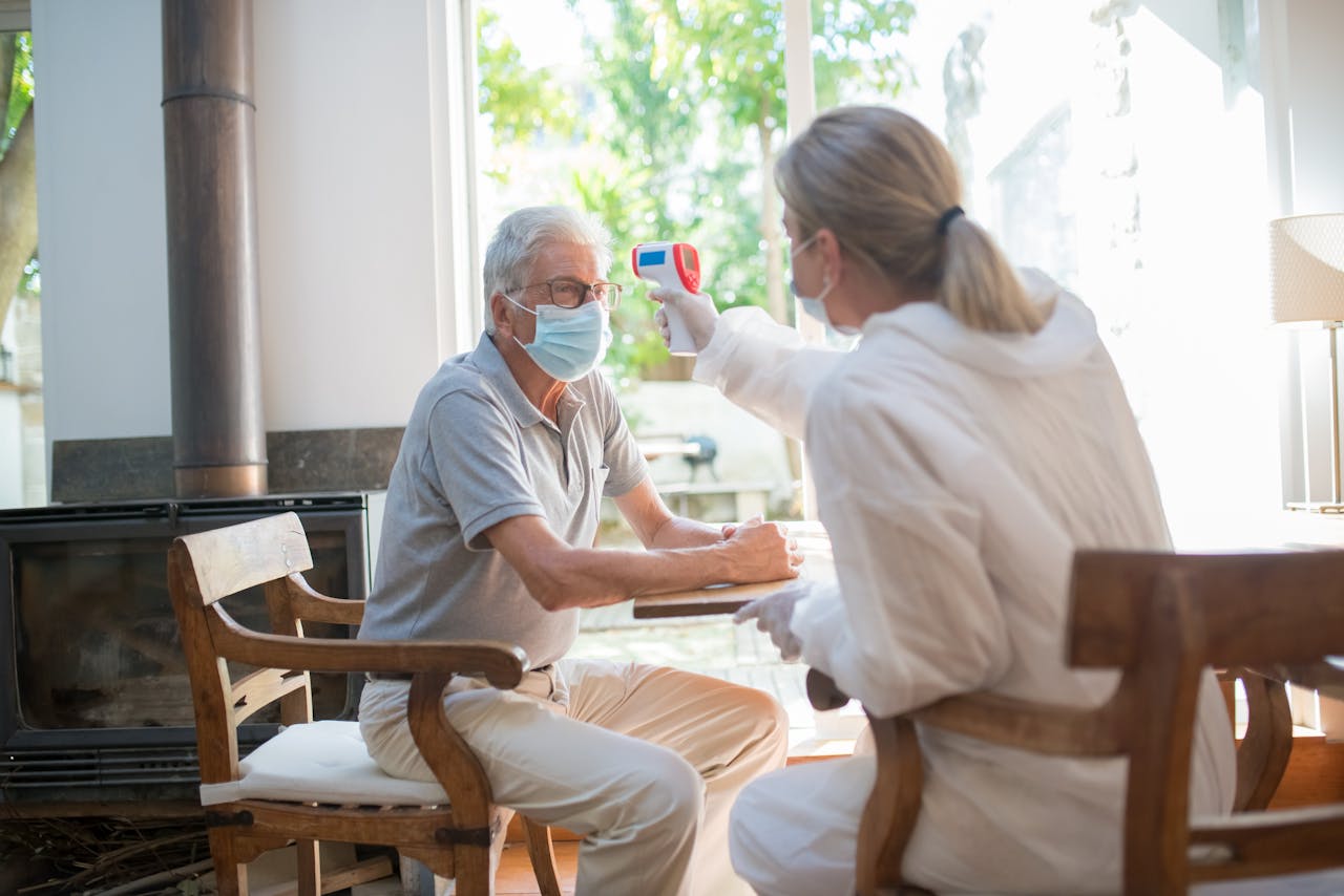Elderly man undergoes temperature check by a healthcare worker in a protective suit and mask indoors.